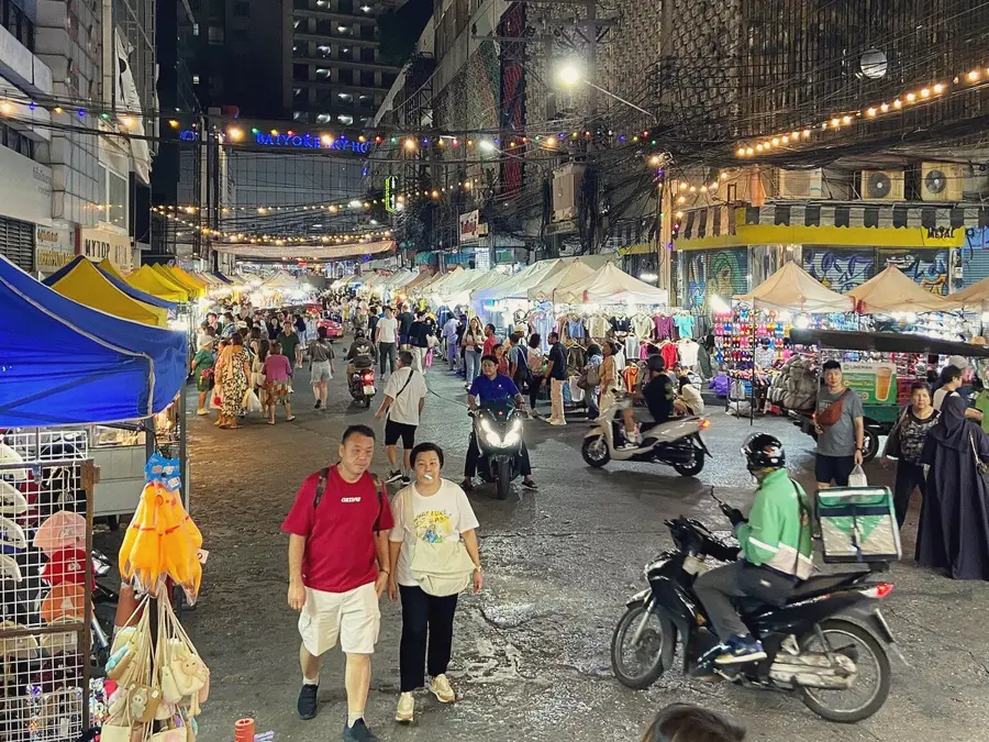 Pratunam night market street lined with lit clothing stalls, Baiyoke Sky Hotel sign in the distance